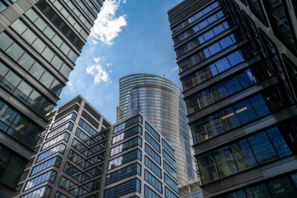 An upward view of a modern skyscraper surrounded by adjacent high-rise office buildings, creating a dramatic urban canyon effect. The central tower's sleek glass design reflects the clear blue sky, while the surrounding buildings enhance the depth and complexity of the scene.