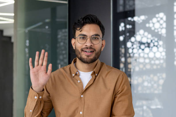 istock Young man on video call waving hand and looking at camera in modern office 2159514855