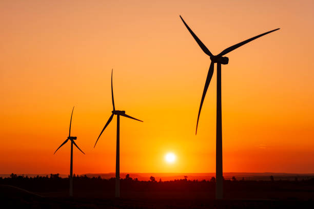 istock Large wind turbines with blades in field on sky background with bright orange sunset. Silhouettes of windmills, big orange sun disk in summer. Alternative energy sources. Ecological energy 2159517093
