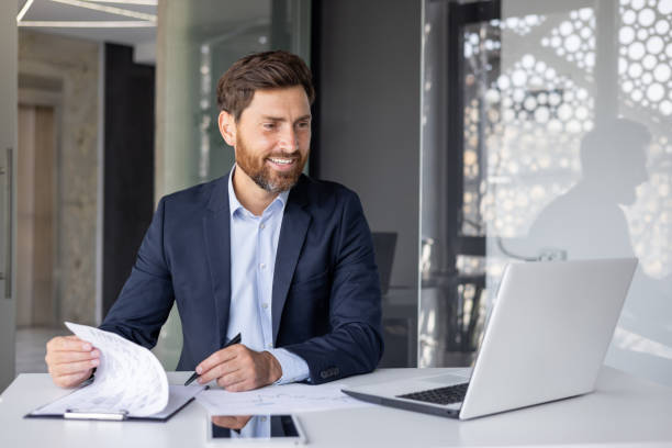 istock Smiling young man working in office with documents, sitting at desktop with laptop and holding online meeting with clients and partners, signing agreement and invoices 2159517497