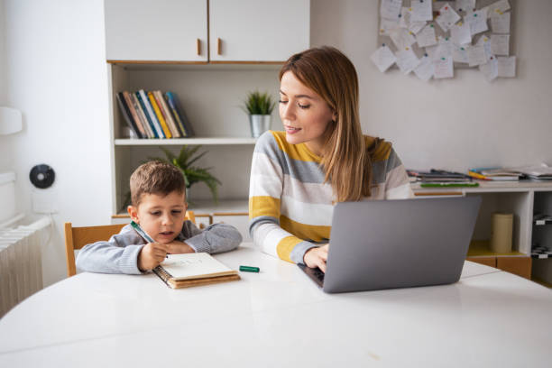 istock Mother using laptop sitting beside son writing in book at home 2159521346