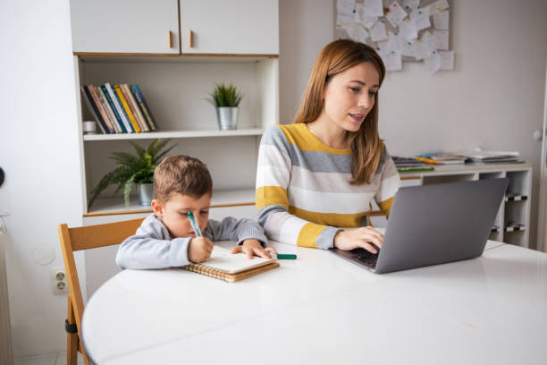 istock Working mother using laptop beside son doing homework at home 2159521421
