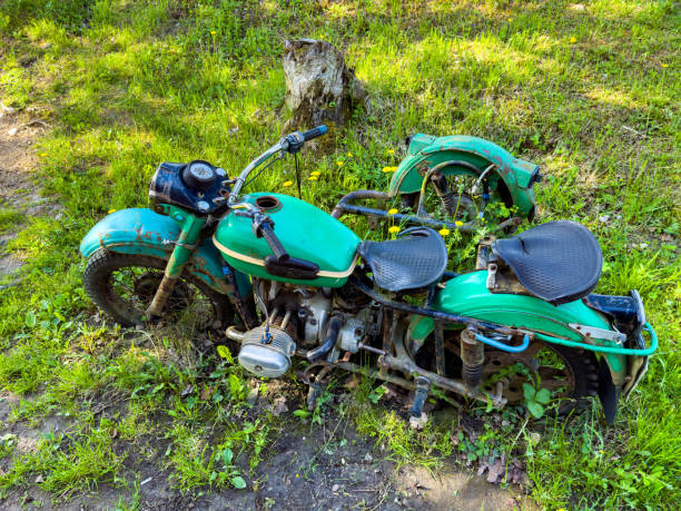 An old, abandoned green motorcycle sits partially buried in overgrown grass. The weathered, rusted parts and vibrant green surroundings create a nostalgic scene, perfect for themes of decay, history, and forgotten treasures.