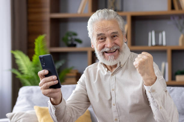 istock Happy senior man celebrating success while using smartphone at home 2159522339