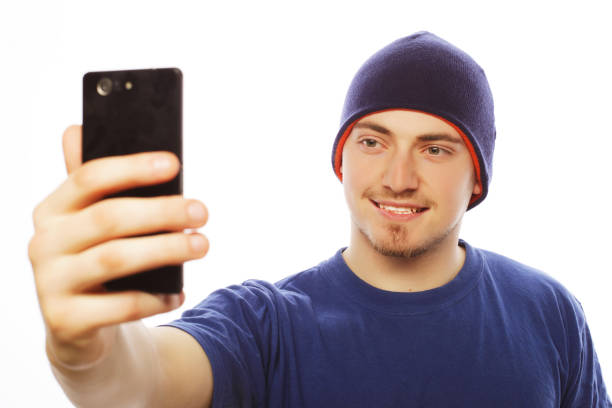 Life style, tehnology and people concept: a young man in shirt holding mobile phone and making photo of himself while standing against white background.