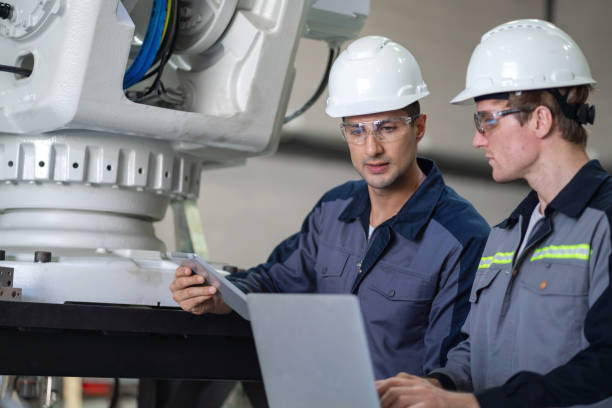 istock Engineers Operating Large Robotic Arm with Remote Controls in Industrial Facility. Two skilled engineers stand in front of a large robotic arm, holding remote controls to operate the machinery while their co-workers enter code into the controller. 2159527240