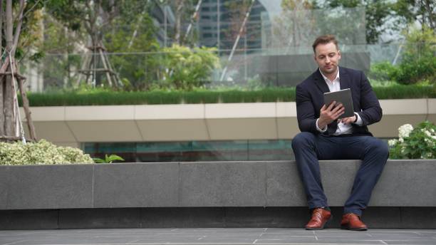 istock Businessman sitting at park while working on tablet by reading report. Urbane 2159527447