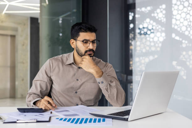 istock A serious young Indian man is sitting in the office at the desk, working with documents, looking thoughtfully at the laptop screen 2159540819