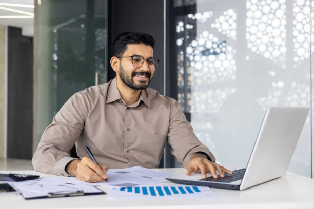istock Smiling businessman working on laptop and analyzing documents at modern office 2159542032