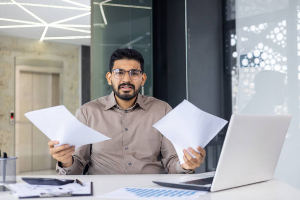 istock Confused businessman holding documents while working at laptop in modern office 2159542121
