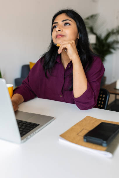 istock Thoughtful indian woman sitting with laptop on table while looking away 2159548993