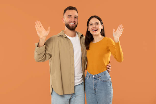 istock Smiling Couple Waving Hands Against Orange Background on Bright Day 2159555741