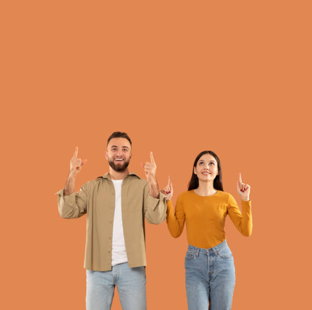 istock Smiling Couple Pointing Upward Against an Orange Background, Blank Space 2159555814