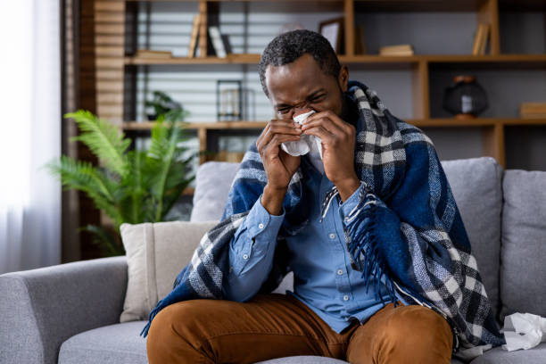 istock Man sneezing and sitting on a sofa with a blanket during a cold at home 2159560106