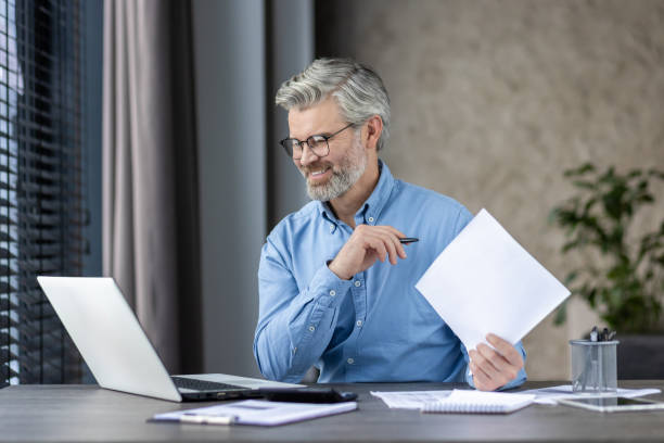 istock A middleaged man is smiling while working on a laptop at his home office, holding papers 2159561833