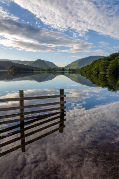 Grasmere in the English Lake District, with mirror-like still water and reflections of clouds and blue sky towards sunset in mid-June on the evening of summer solstice on a beautiful, calm night, looking towards the surrounding woodland and fells. grasmere en el distrito de los lagos ingleses, con agua tranquila como un espejo y reflejos de nubes y cielo azul hacia el atardecer a mediados de junio en la tarde del solsticio de verano en una noche hermosa y tranquila, mirando hacia el bosque y las col