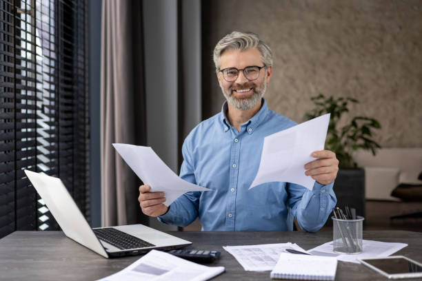 istock Smiling businessman working with documents at desk with laptop in modern office 2159564661