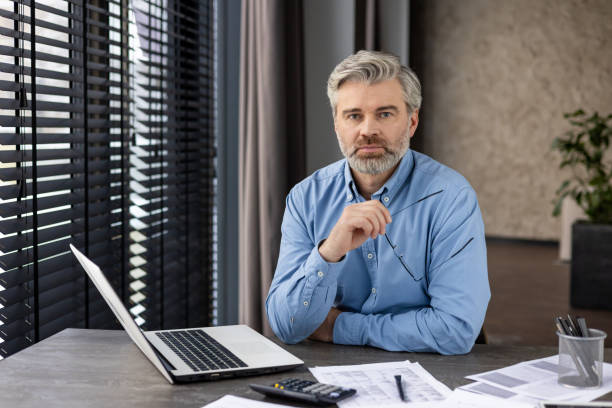 istock Confident businessman at desk holding glasses, working on laptop, and analyzing documents 2159567970
