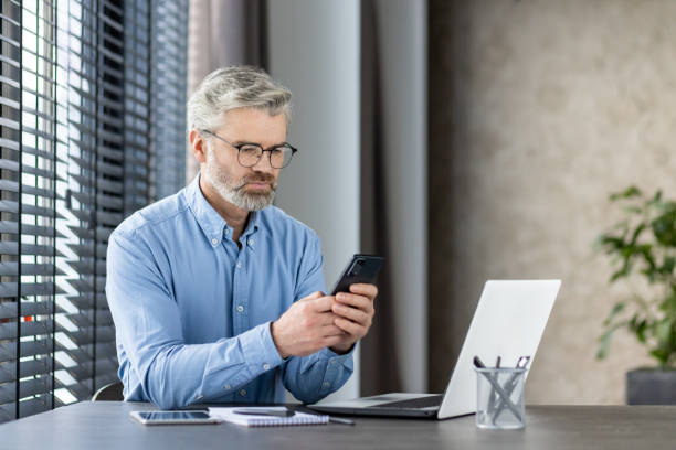 istock Mature businessman focused on mobile phone while working on laptop at office desk 2159572345