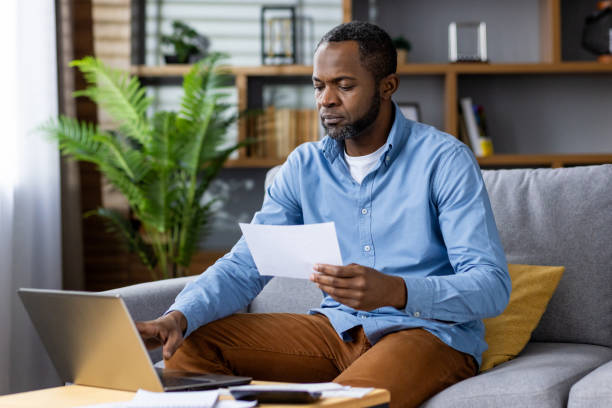 istock A man is working from home, reviewing a document and using a laptop in his cozy living room, portraying a modern workspace, focusing on his task with dedication and efficiency 2159574456