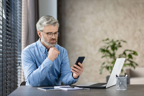 istock Focused mature businessman using smartphone while working on laptop in office 2159574874
