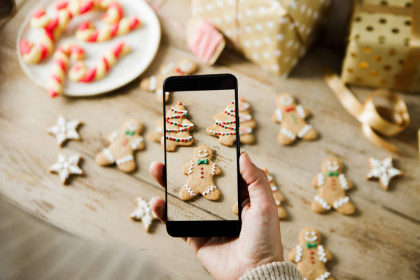 Woman hands photographing beautifully decorated Christmas gingerbread cookies on wooden table with candie canes and gift boxes. Various types of a Christmas cookies on kitchen counter being photographed using phone.