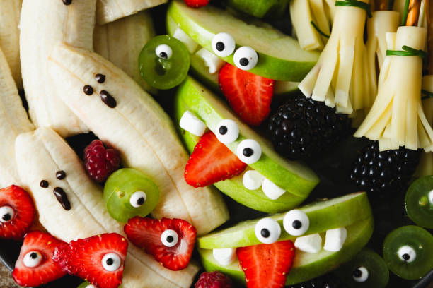 Close-up of spooky banana ghosts monsters, strawberry and green apple monsters for Halloween party. Halloween party fruit serving display. Festive Halloween table setup with funny decorated fruits.