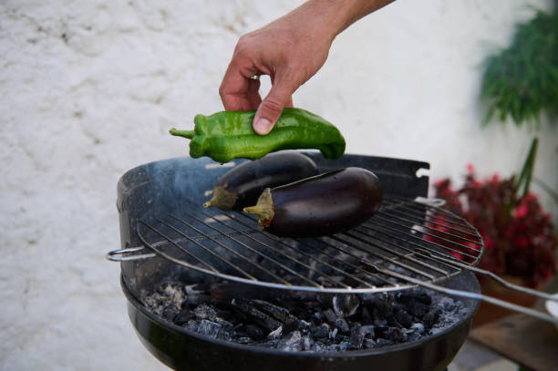 A hand placing a green pepper on a barbecue grill with two eggplants, showcasing outdoor cooking and healthy eating.