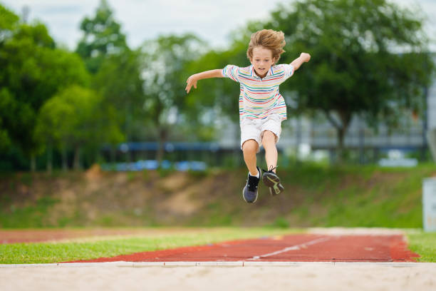 A group of children participating in an outdoor athletics event, practicing long jump on a sports field. The activity emphasizes teamwork, motor skills, and enjoying physical exercise in a fun environment.
