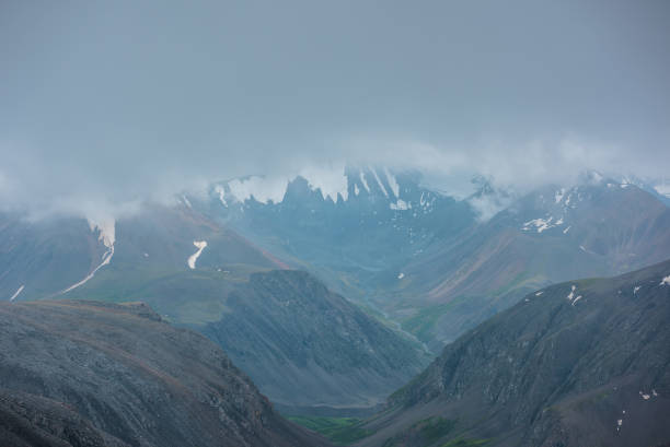 istock Large snow mountain range silhouette in rainy low clouds under gray sky. Mountain stream in green valley in light transparent mist. Hills and rocks in bad weather. Low cloud on big snowy mountains. 2159601147