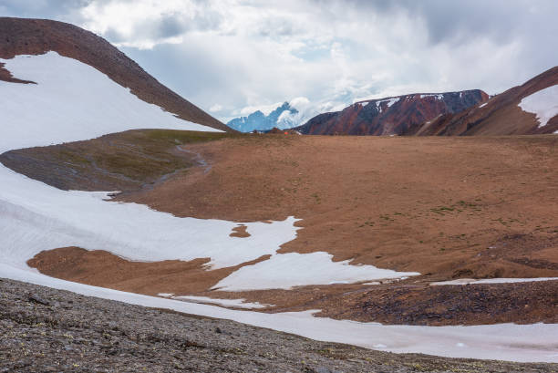 High alpine valley with sunlit glacier against large pyramid shaped pointy peak in low clouds. Giant mountain peaked top of triangle shape far away. Scenic view to pinnacle silhouette in cloudy sky.