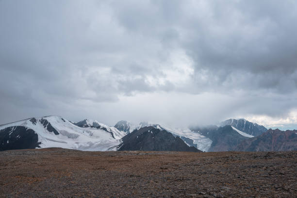istock Dramatic misty top view from stone pass to big glacier tongue among sharp rocks and large snow-capped mountains in rainy low clouds. Dark atmospheric mountain silhouettes in rain in gray cloudy sky. 2159601157