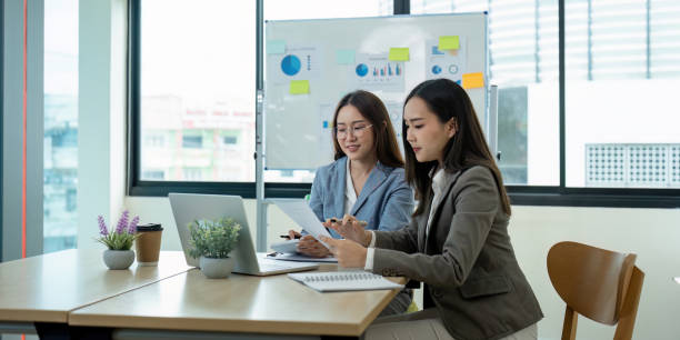 istock Two Businesswomen Collaborating in Modern Office with Laptops and Documents, Analyzing Data on Whiteboard 2159612769