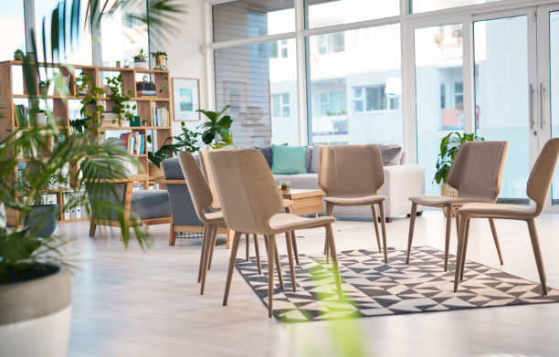 A modern support group meeting room featuring comfortable chairs arranged in a circle, natural light, and lush indoor plants. The space is designed to foster open communication and a sense of community.