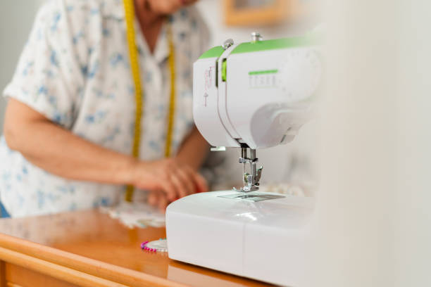 A senior woman in the background, using chalk to mark fabric, with a modern sewing machine prominently in the foreground.