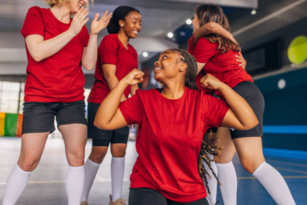 A joyous celebration unfolds as the women's soccer team celebrates their victory with hugs and cheers indoors, all dressed in red shirts and black shorts with white socks.