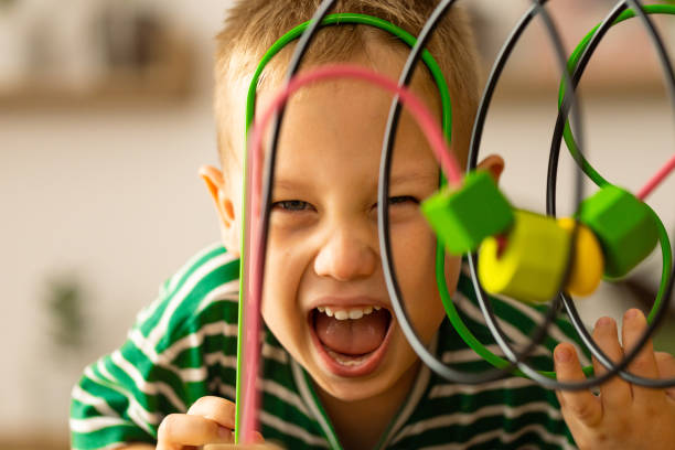 Caucasian boy is playing with a wooden developmental toys on table at home. Boy makes fanny faces  and smiling.