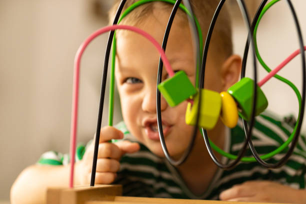 Caucasian boy is playing with a wooden developmental toys on table at home. Boy makes fanny faces  and smiling.