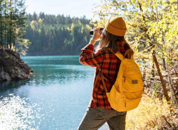istock Young woman with a yellow backpack looking through binoculars at waterfowl in autumn on a lake Birdwatching and eco tourism 2159684020