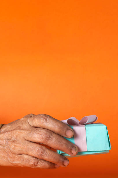 istock Senior Woman's Hand Holding Gift Box on an Orange Background 2159685673