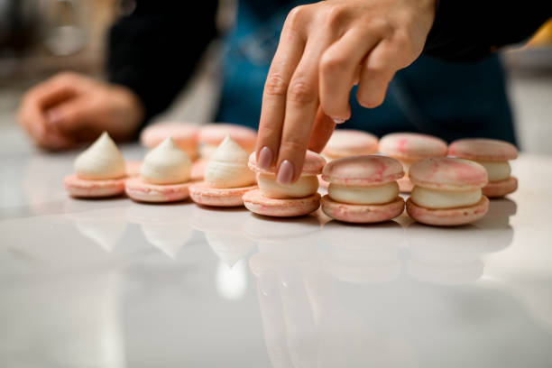 istock Fair skin chef's hands makes a pink and beige macarons on a white marble table 2159695409