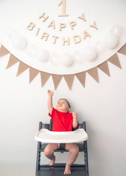 A one-year-old baby sits in a baby chair against the backdrop of a birthday arrangement for his first birthday photo