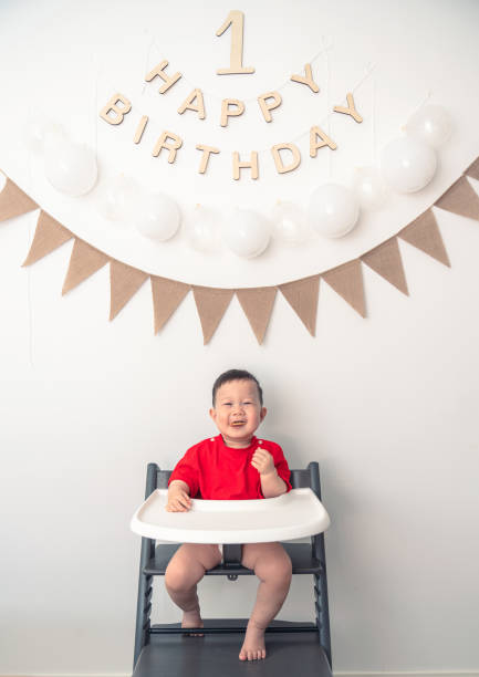 A one-year-old baby sits in a baby chair against the backdrop of a birthday arrangement for his first birthday photo