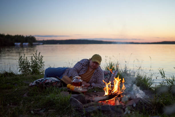 a man sits on a river bank on a summer evening at sunset next to a fire and drinks tea