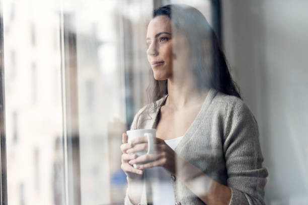 istock Attractive woman drinking a cup of coffee while looking forward standing next to the window at home. 2159719241