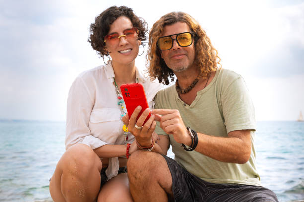 istock Smiling couple on the beach browsing smartphone apps looking at camera 2159766961
