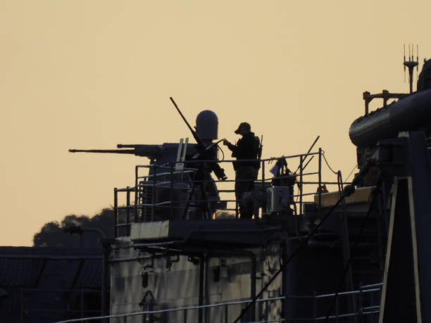 An armed crew member checks the ammunition box of a 0.5 calibre machine gun during his patrol of USS Emory S. Land, a submarine tender of the US Navy docked at Garden Island naval base in Sydney Harbour. In the background, with the long barrel and radar dome is a MK 38 machine gun system. This image was taken on a cold and sunny morning at sunrise on 29 June 2024.