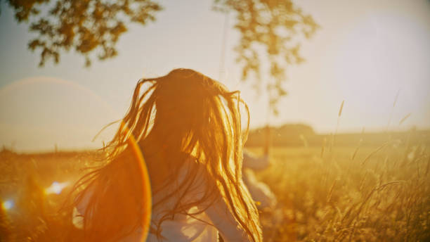 A joyful young girl sprints towards a swing hanging from a solitary tree, her playful activity illuminated by the golden hues of sunset.
