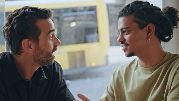 Joyful men talking coffeehouse smiling to each other close up. Cheerful two multiethnic guys fascinated by conversation in cafeteria. Couple diverse colleagues enjoy friendly communication in cafe.