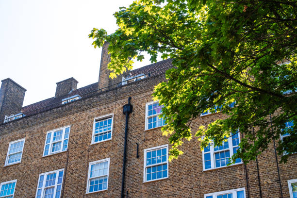 Typical red brick townhouses in London. The apartments have white framed widows and small French balconies. In front of building there are some green bushes.